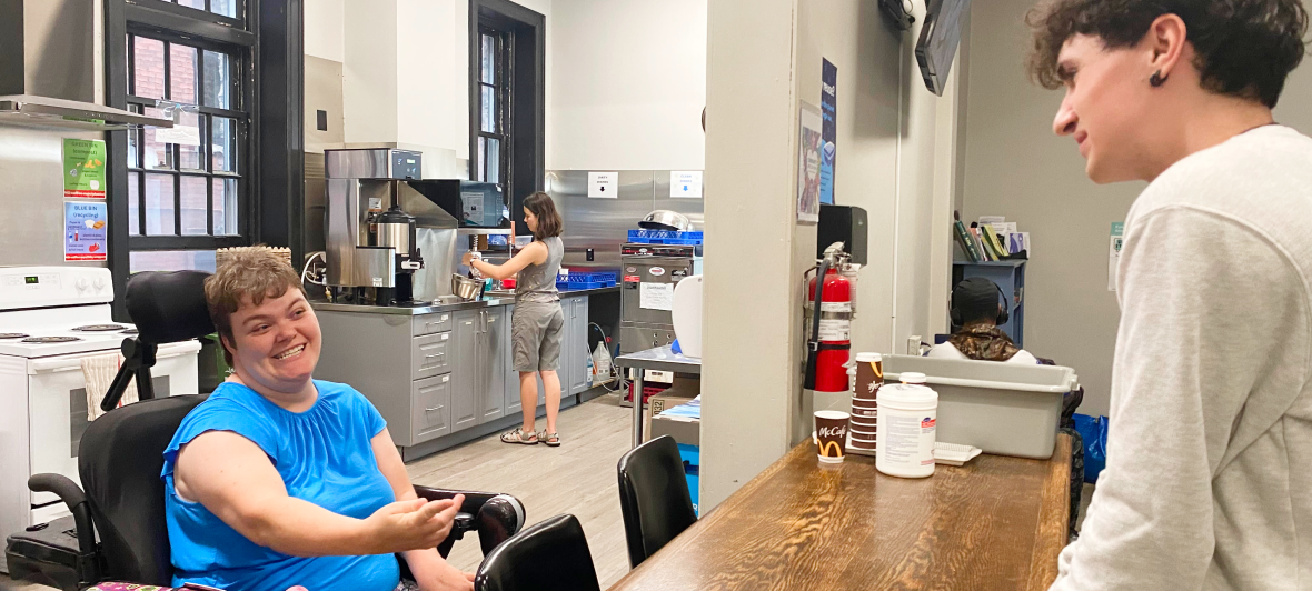 A smiling young woman in a wheelchair serves a young customer who is standing at a café counter.